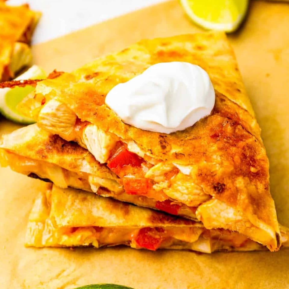 Stack of golden-brown quesadilla wedges filled with chicken and red peppers, topped with sour cream and lime.