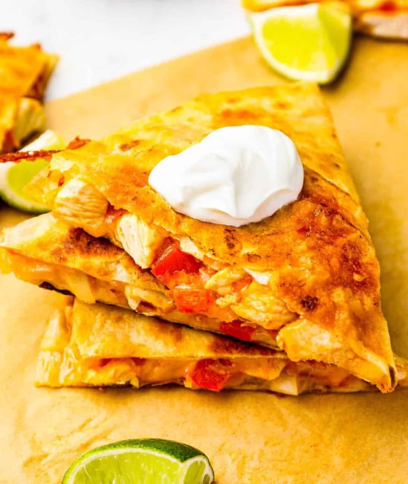 Stack of golden-brown quesadilla wedges filled with chicken and red peppers, topped with sour cream and lime.