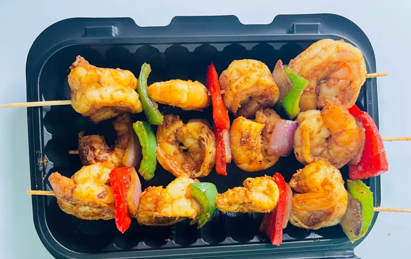 Three Caribbean shrimp and vegetable skewers with red and green peppers, and onions in a black container.